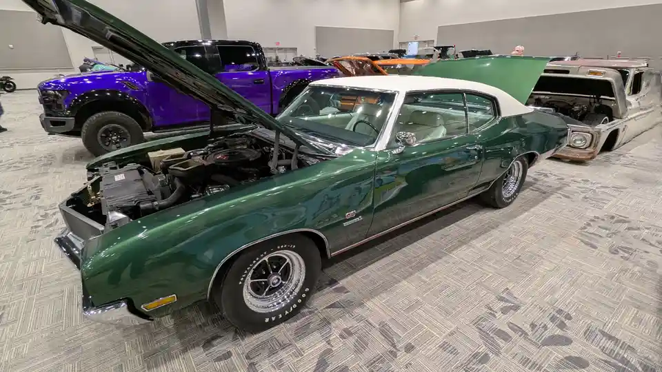 A green classic muscle car with its hood open is displayed at an indoor car show, surrounded by other vehicles including a…