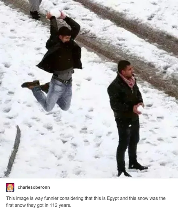 Two men are in a snowy outdoor setting, one is mid-air throwing a snowball with arms raised, while the other stands nearby…