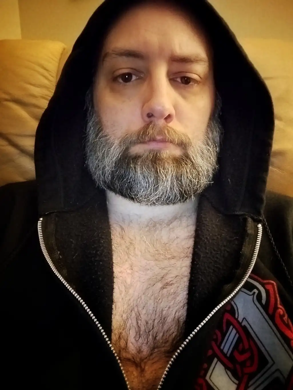 a man with a gray beard and mustache, wearing a black zip-up hoodie with the hood up, sits on a beige couch. His zipper is…
