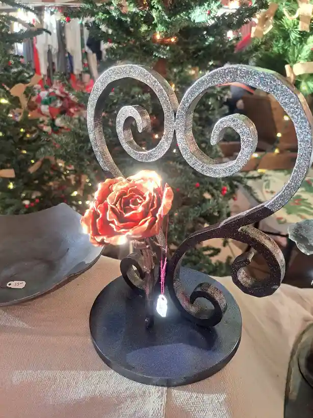 A decorative metal sculpture shaped like a heart with a glowing red rose at its center sits on a table in front of a…