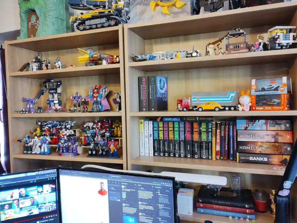 A wooden bookshelf is densely packed with collectibles including numerous Transformers action figures, large Lego models…