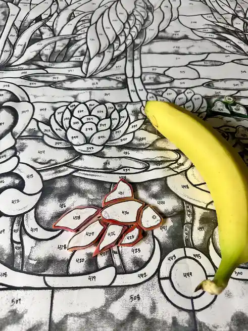 A yellow banana is placed on a black-and-white coloring book page featuring a lotus flower and other intricate patterns…