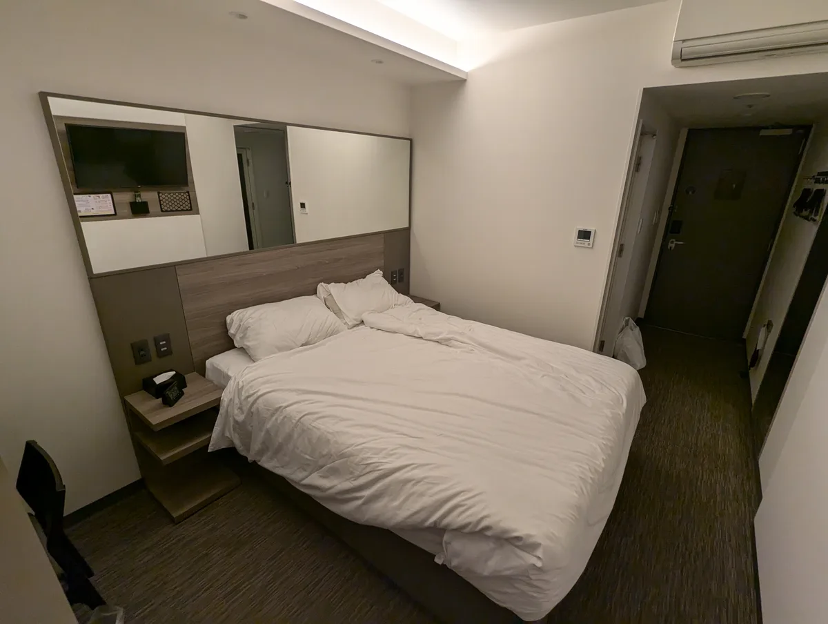 A narrow, modern hotel room with a large bed covered in white linens, a wall-mounted TV reflected in a large mirror above…