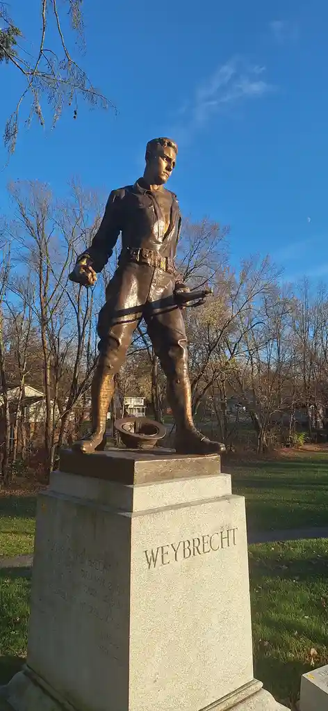 A bronze statue of a soldier stands on a stone pedestal in a park with bare trees and a blue sky; the pedestal is engraved…