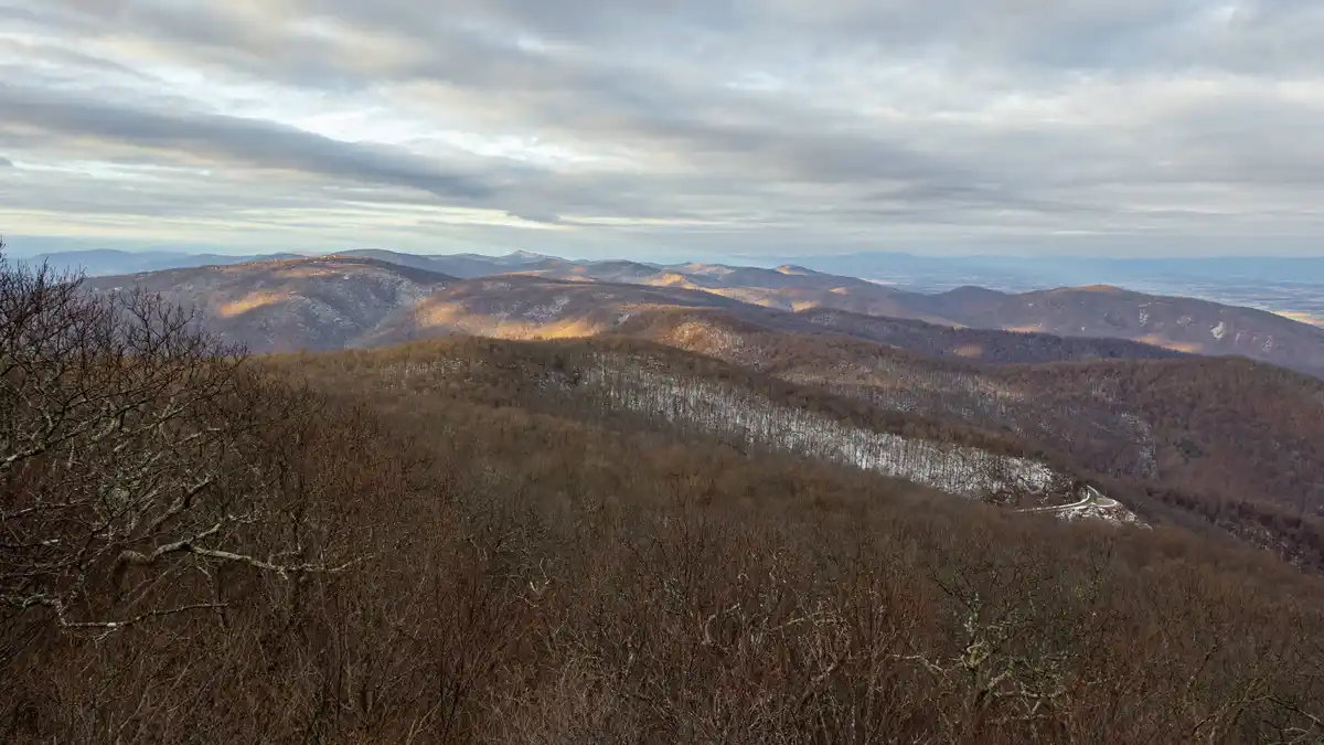 A wide landscape photo of rolling, snow-dusted mountains under a cloudy sky, with bare winter trees in the foreground and a…
