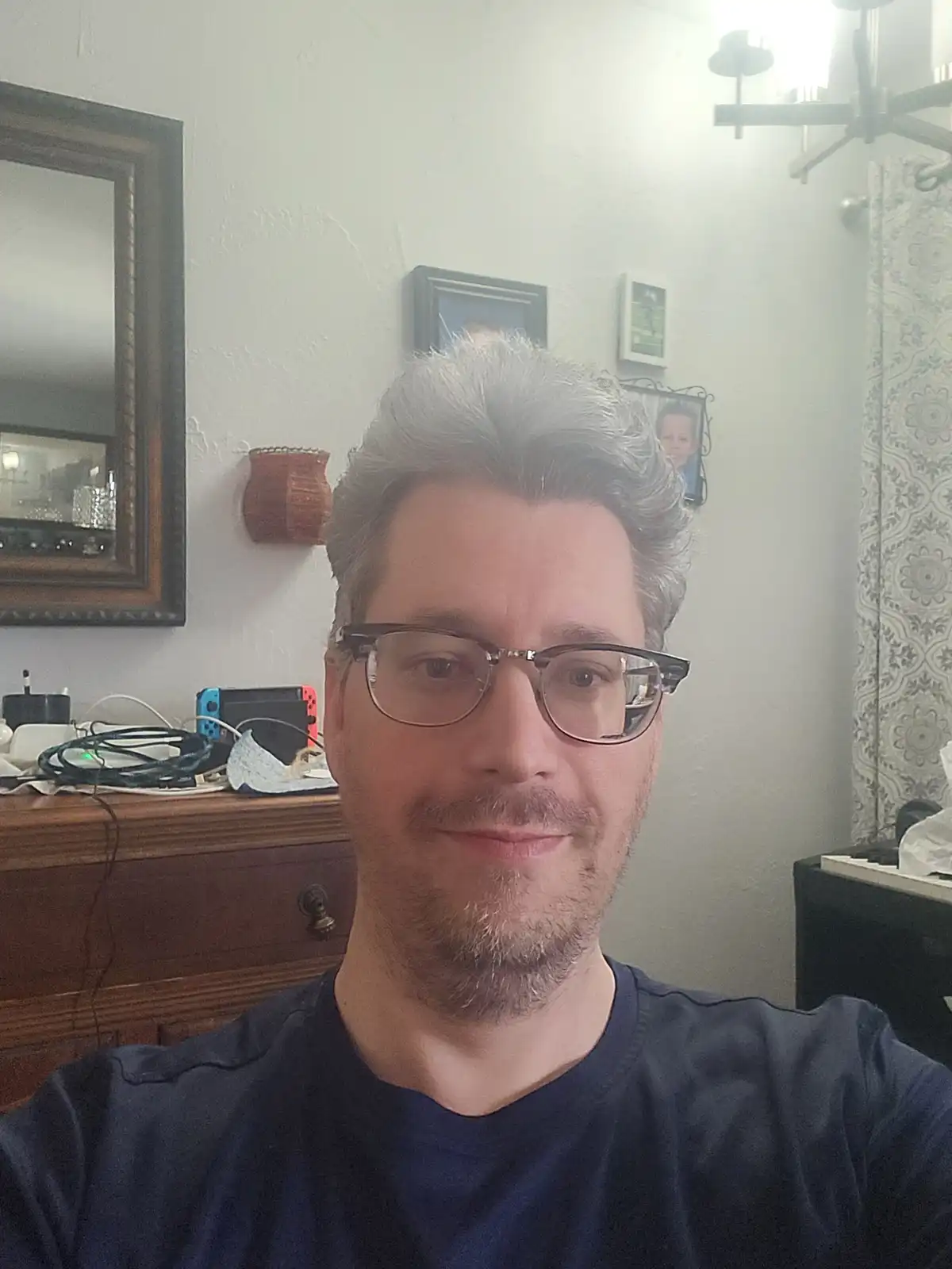 A selfie of a man with graying hair and glasses, smiling slightly while wearing a dark blue t-shirt. Behind him is a wooden…