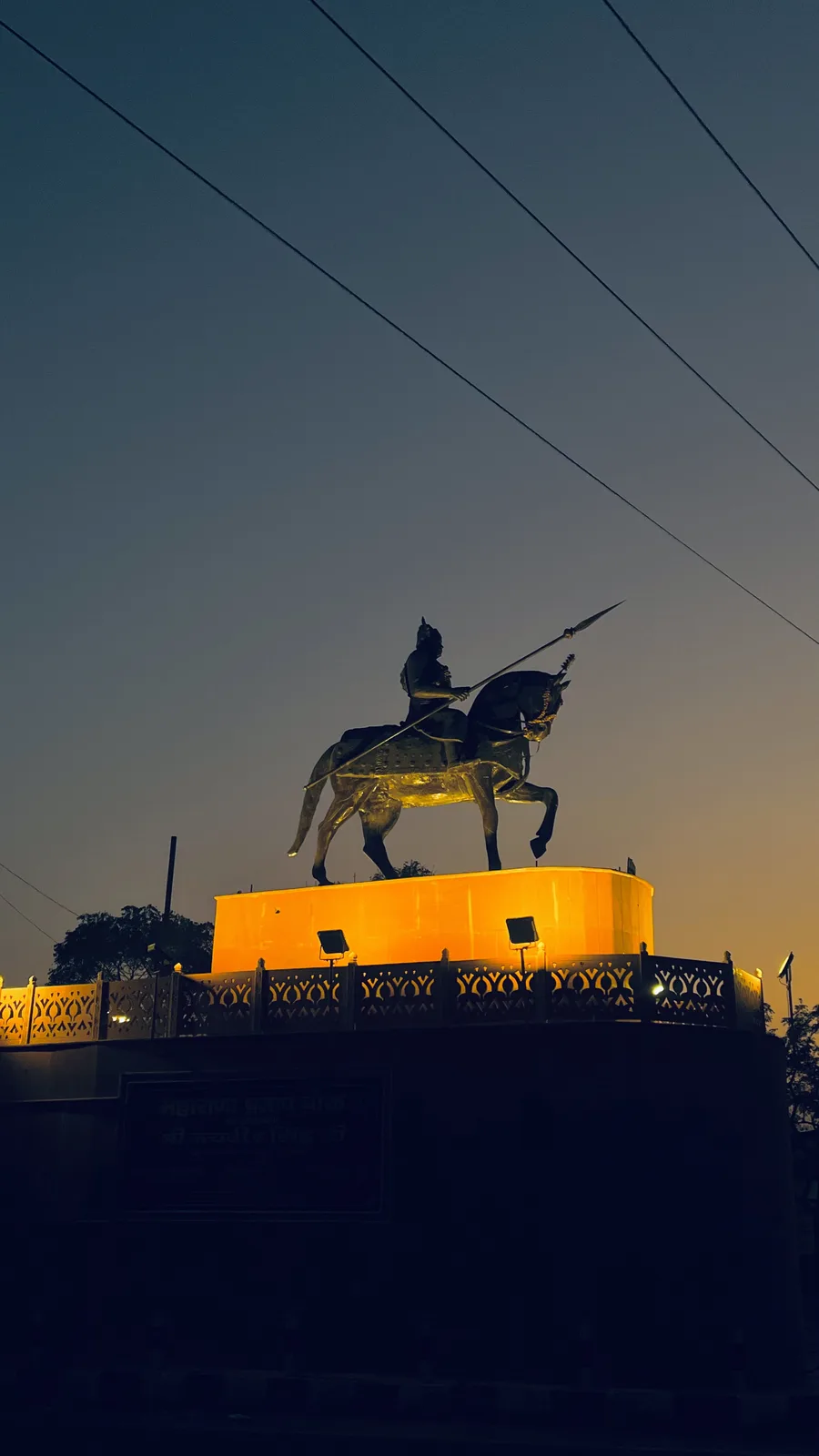 A low-angle photo at dusk shows a large, illuminated equestrian statue of a warrior holding a spear, standing atop a…