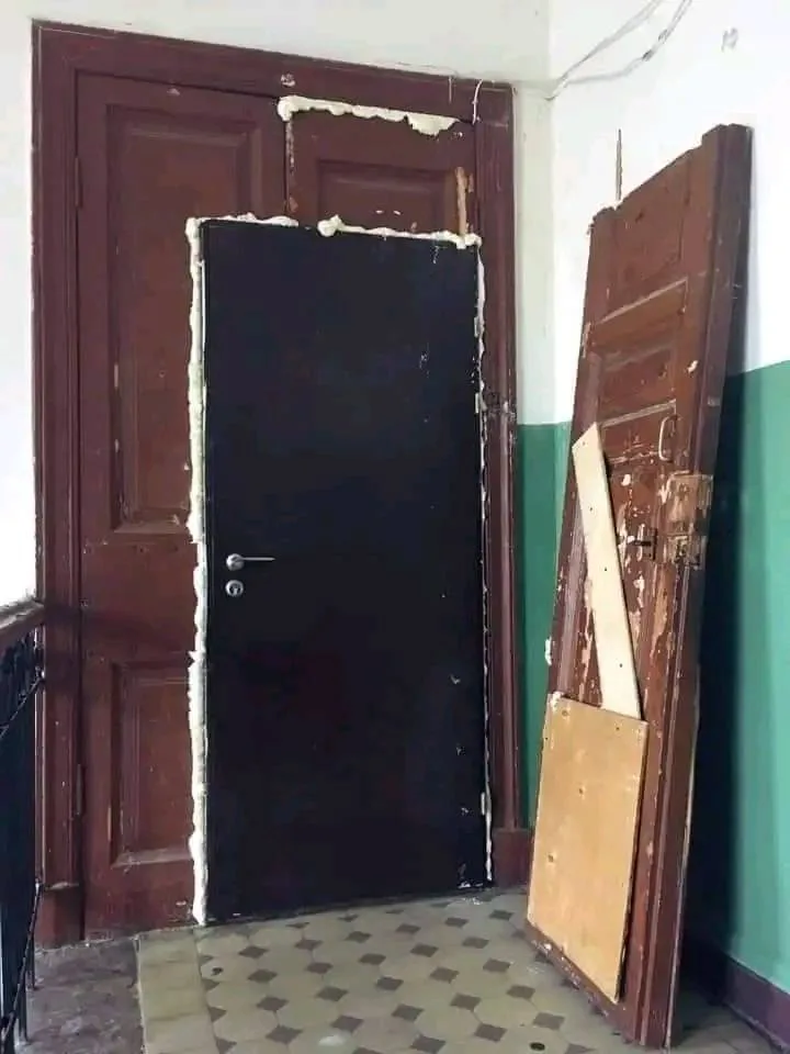 A dark, plain door is set into a wooden door frame with thick white foam sealant around its edges. To the right, an old…