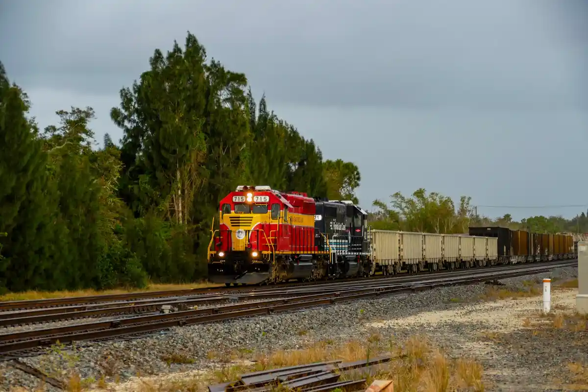A red and yellow Florida East Coast locomotive, numbered 715, pulls a long freight train through a wooded area under an…