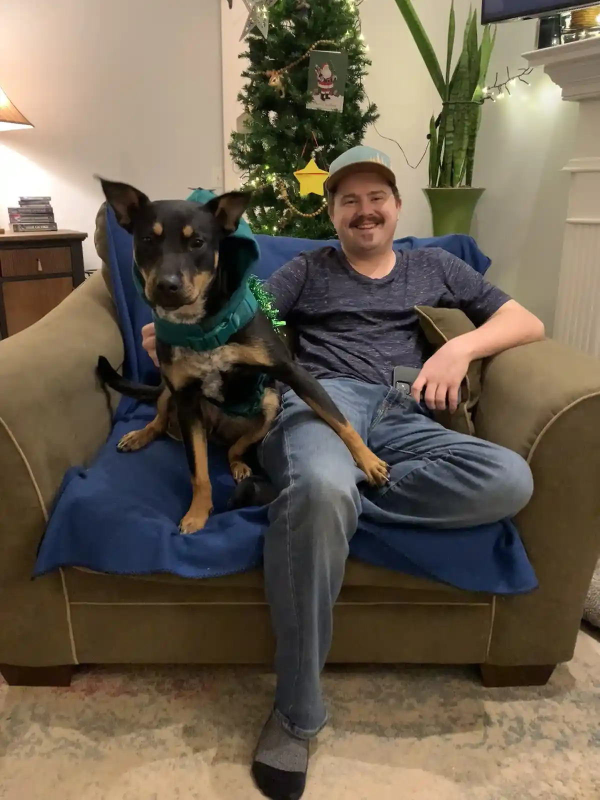A man with a mustache and cap sits on a couch next to a dog wearing a teal hooded costume, both looking at the camera…