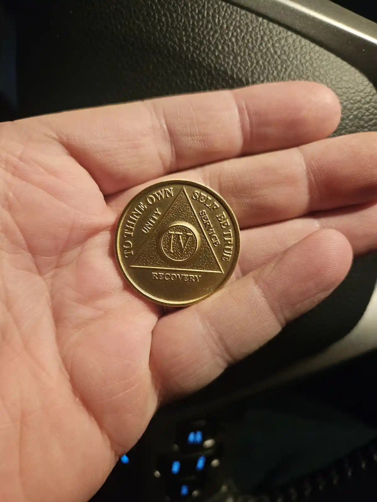 A hand holds a gold-colored recovery token featuring a triangle with the words 'to think own unity', 'self service', and…