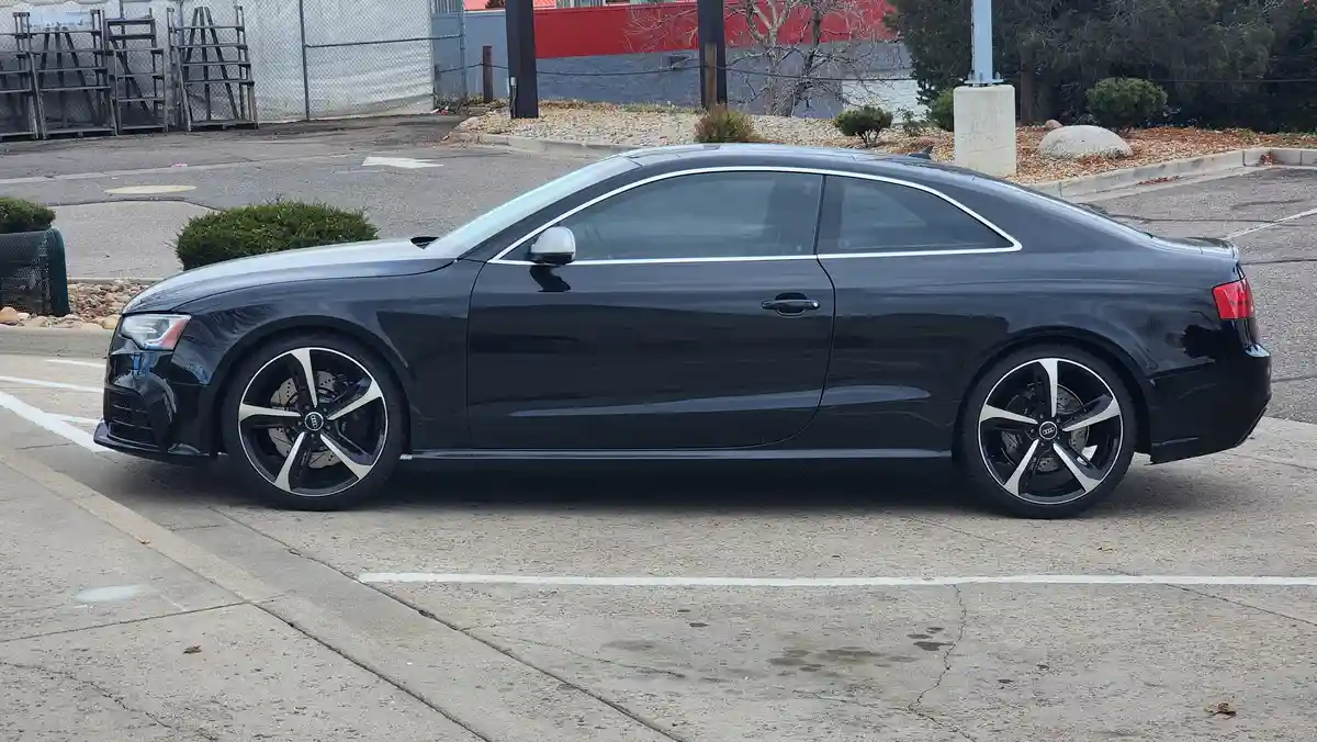 A black Audi coupe is parked in a lot, showing its sleek side profile with silver trim and alloy wheels. The overlaid text…
