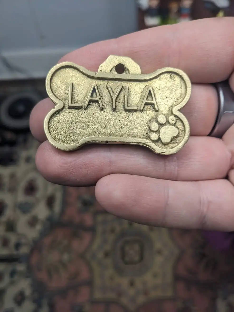 A close-up photo shows a person's hand holding a gold-colored, bone-shaped pet ID tag. The tag has the name 'LAYLA' embossed…