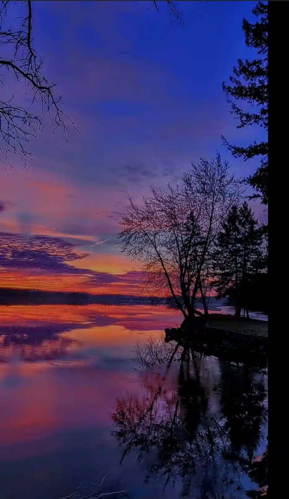 A serene landscape photo of a lake at sunset, with vibrant orange and purple clouds reflected in calm water, framed by…