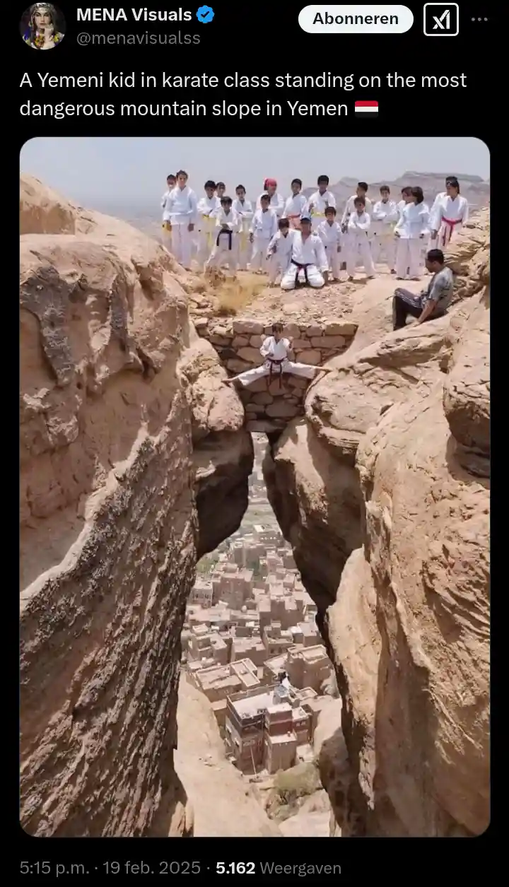 A young boy in a white karate gi performs a split on a narrow stone ledge high above a valley town, with other children in…