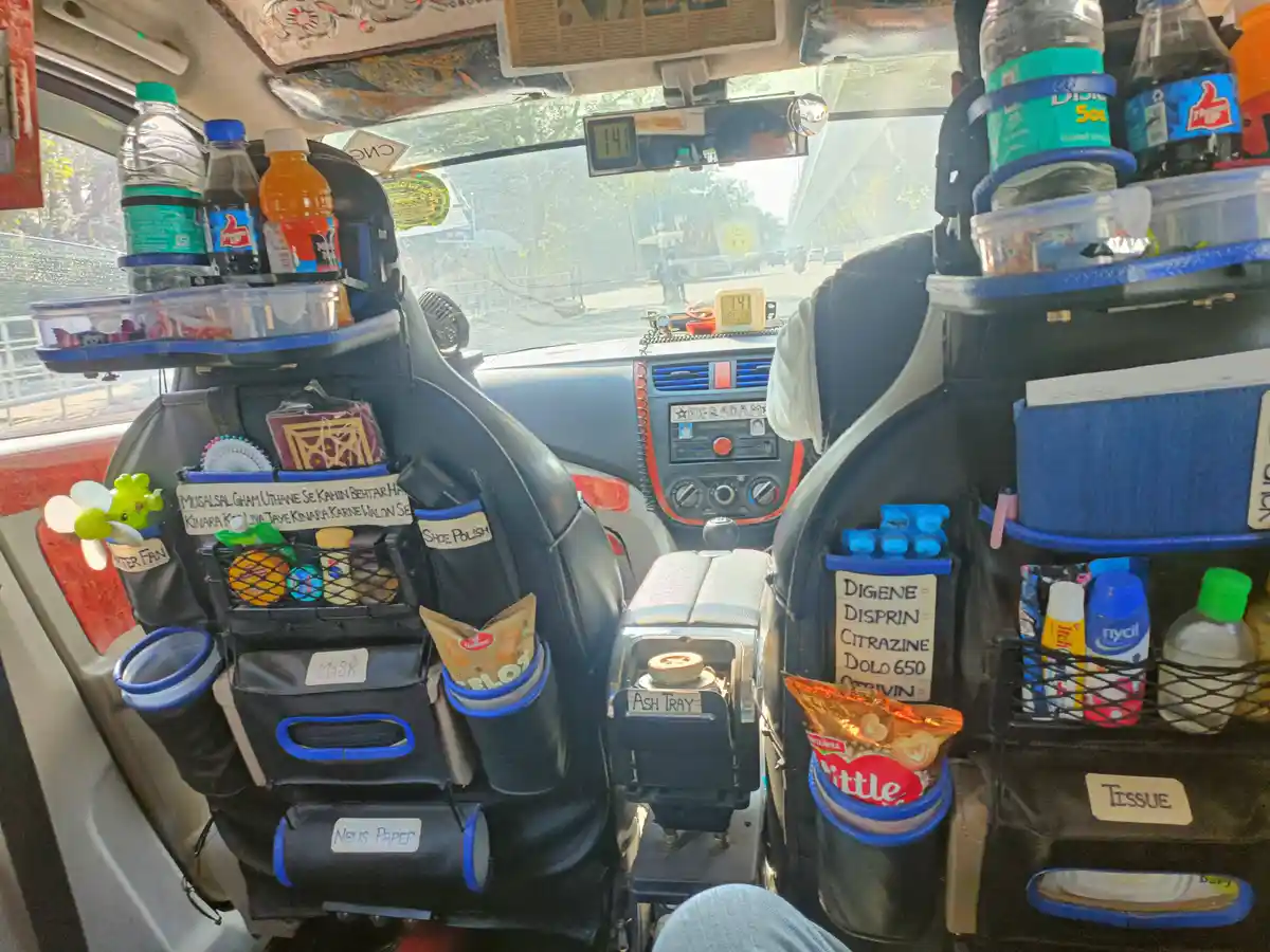 The interior of a heavily customized auto-rickshaw, with both front seats covered in black organizers holding bottles…