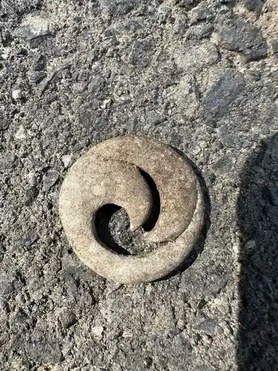 A close-up photo shows a weathered, spiral-shaped stone object lying on rough asphalt pavement. The stone has a distinct…