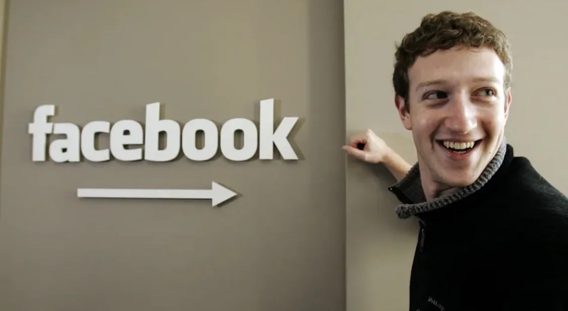 A man with curly brown hair and a black jacket smiles while peeking from behind a wall with a white 'facebook' logo and…