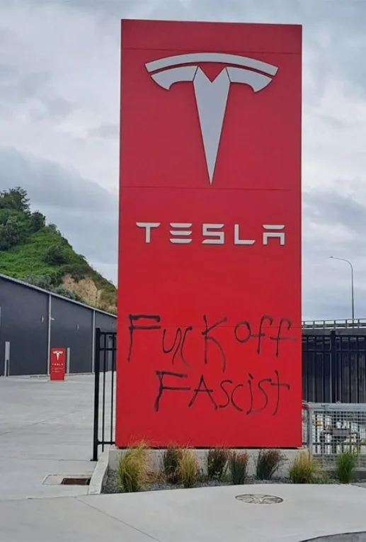 A large red Tesla sign with the company logo and name is visibly defaced with black spray-painted graffiti that reads 'Fuck…