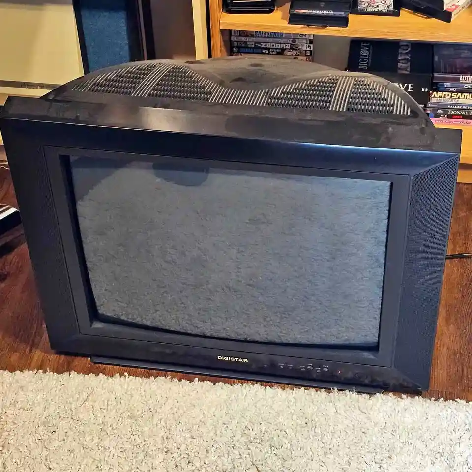 A dusty, old black CRT television with a 'DIGISTAR' brand name sits on a white carpet, its screen showing static. Behind it…