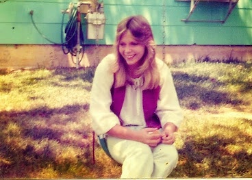 A woman with long blonde hair, wearing a white blouse and a purple vest, sits on a chair outdoors in front of a teal wall…
