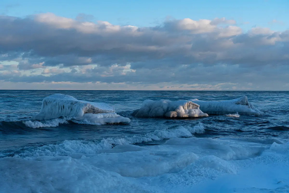 A serene winter seascape shows large, jagged ice formations floating in dark blue water under a cloudy sky with soft pink…