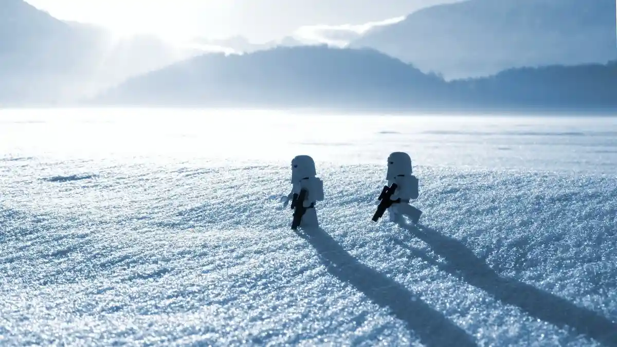 Two LEGO Star Wars snowtrooper figures stand in a vast, sunlit snowy field with mountains in the hazy background. The…