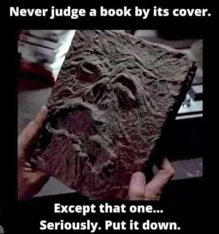 A person holds a book with a cover that is clearly shaped like a grotesque, screaming face. The top text reads 'Never judge…