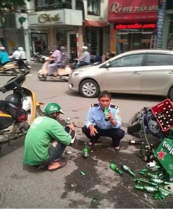 Two men are crouched on a busy street amid spilled beer bottles and broken glass. One man wears a green Grab delivery helmet…