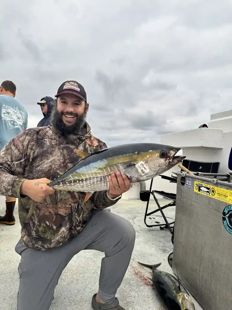 A bearded man in a camouflage jacket and cap kneels on a boat deck, proudly holding a large tuna fish with a tag labeled…