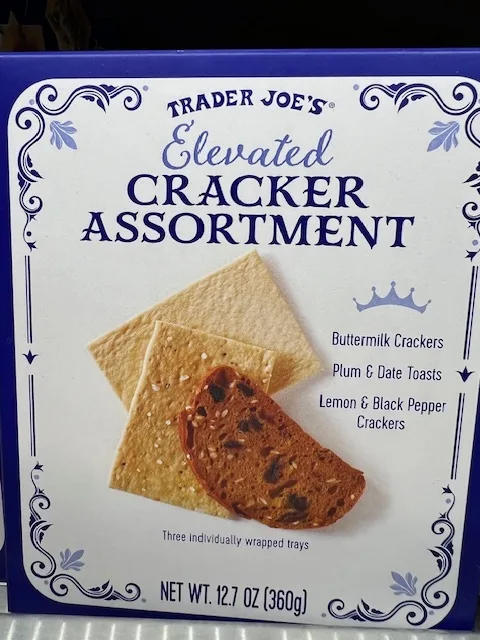 A close-up of a Trader Joe's 'Elevated Cracker Assortment' box, featuring images of three types of crackers: Buttermilk…