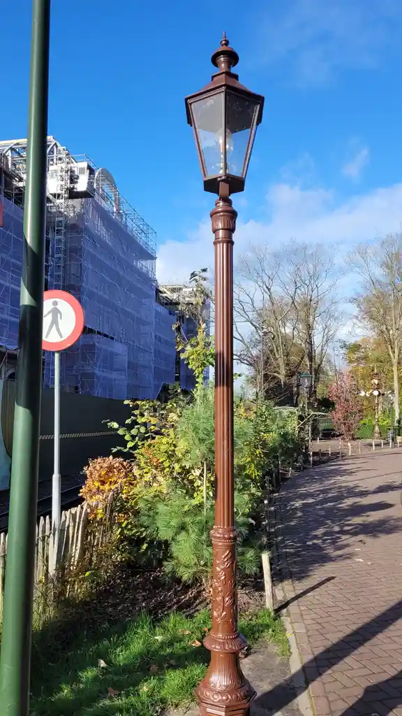 A brown ornate street lamp stands on a brick sidewalk next to greenery, with a 'no walking' pedestrian sign visible nearby…