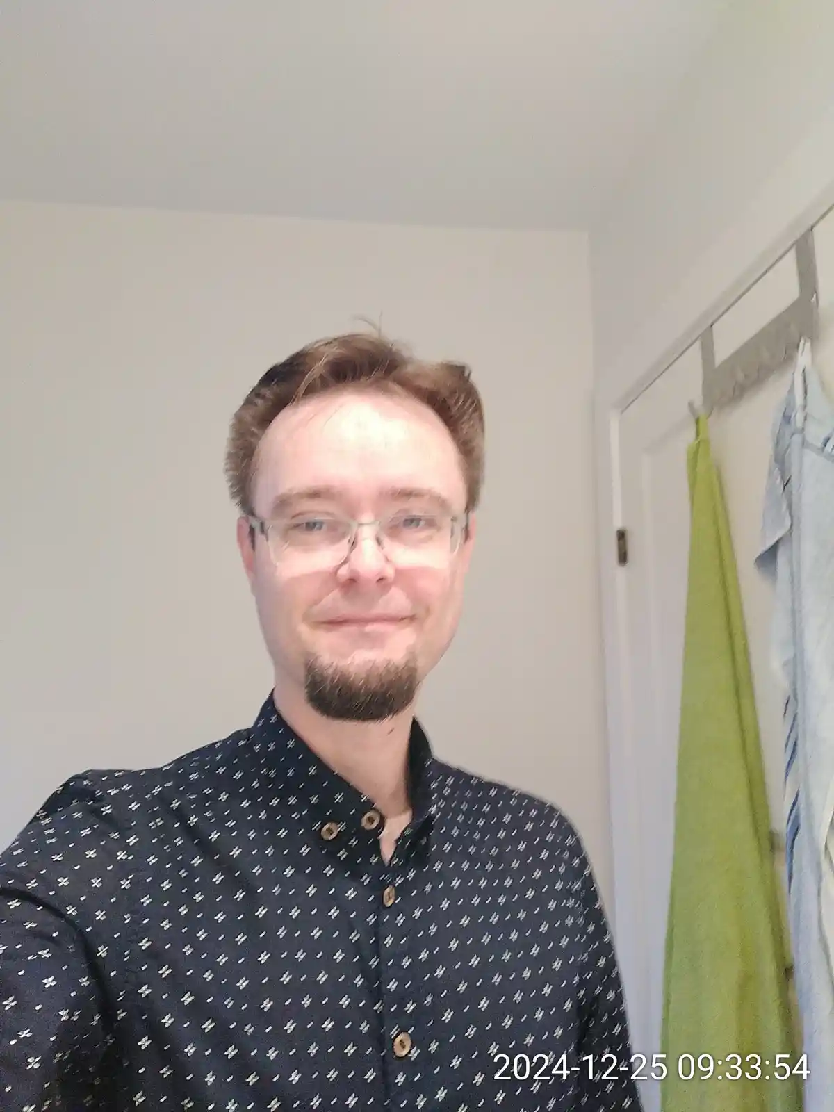 A selfie of a man with glasses and a goatee wearing a dark patterned button-up shirt, standing in a room with a white wall…