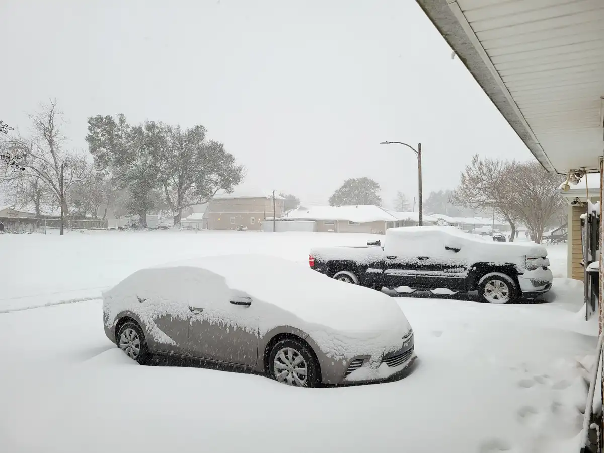 A heavy snowfall blankets a residential driveway, completely covering a gray sedan and a black pickup truck parked side by…