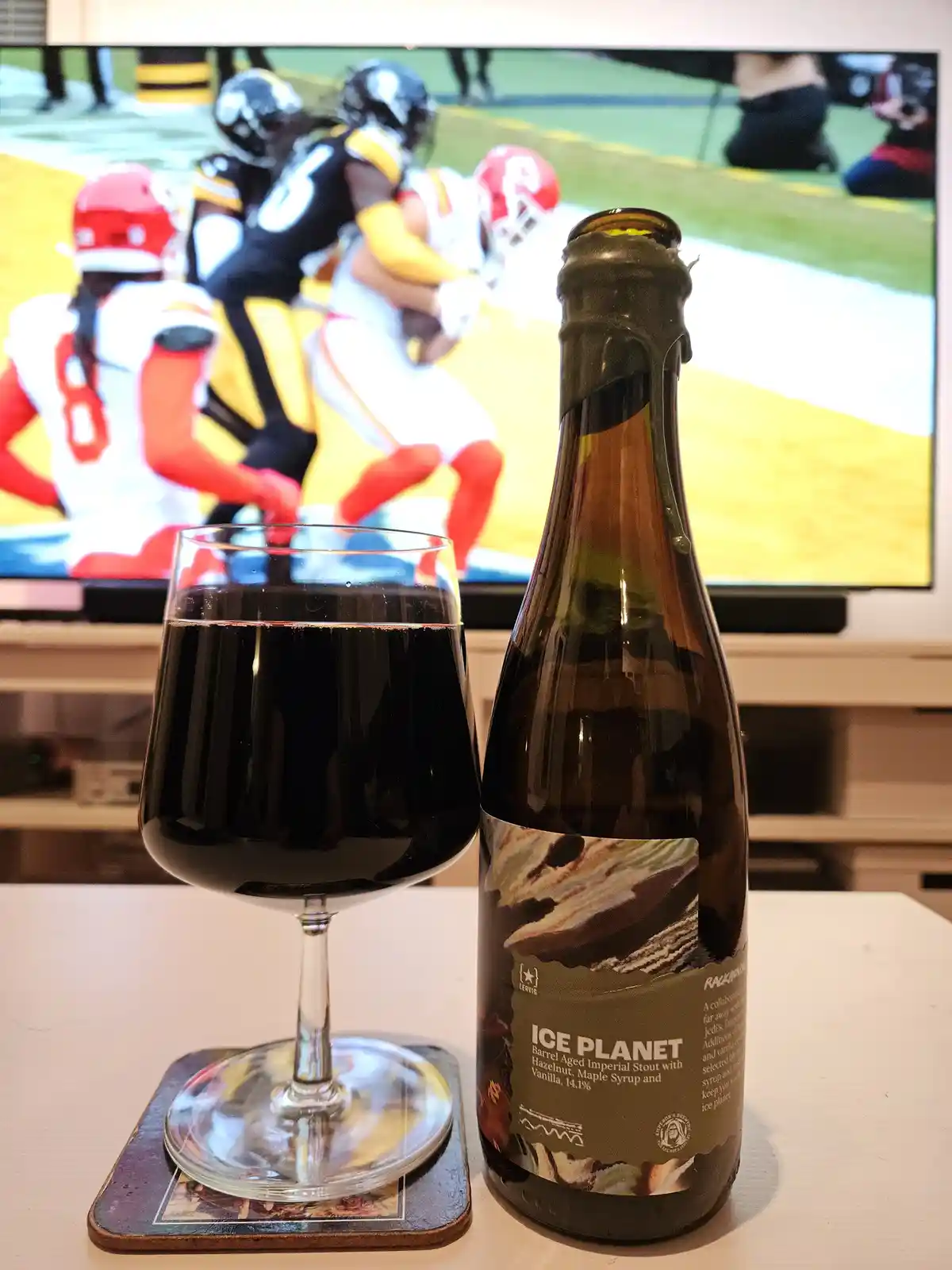 A bottle of 'Ice Planet' beer and a glass of dark liquid sit on a table in front of a TV showing an American football game…