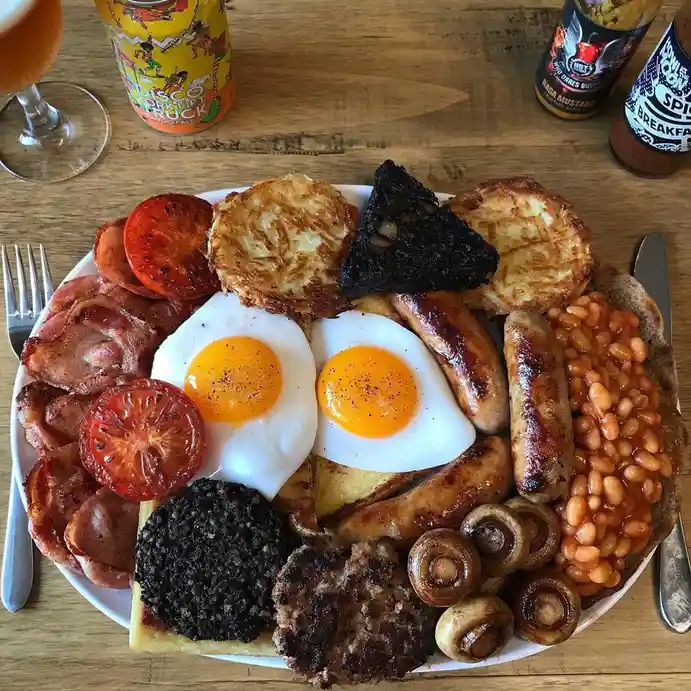 A top-down photo of a very full English breakfast plate with fried eggs, bacon, sausages, baked beans, mushrooms, black…