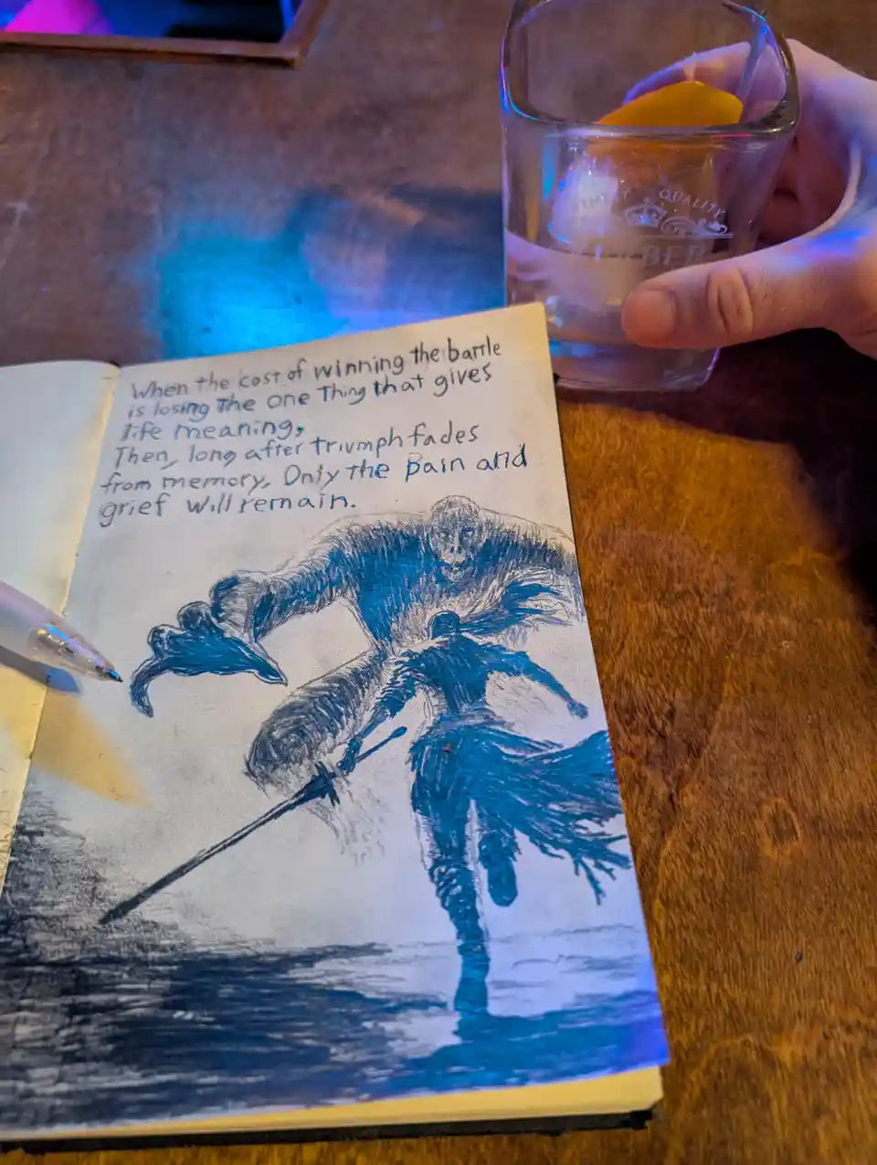 A hand holds a glass with an orange slice while a notebook on a wooden table displays a blue ink drawing of a warrior…