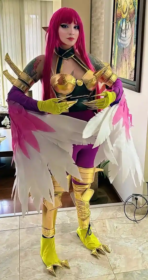 A person in a detailed cosplay of a fantasy character with bright pink hair, gold and purple armor, and large white and pink…