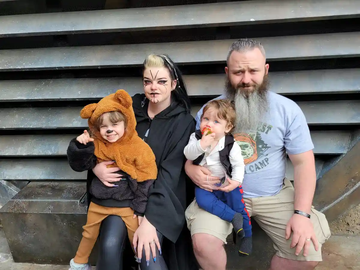 A family of four poses for a photo: a woman with black face paint and a black robe, a man with a long gray beard in a…