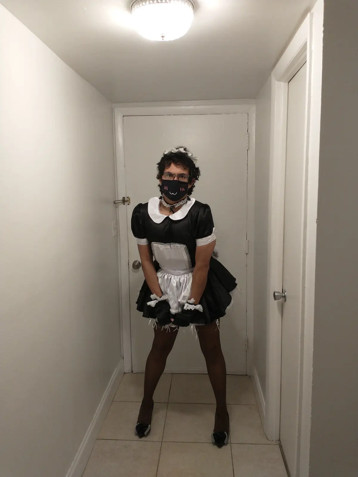 A person with curly hair and glasses stands in a narrow hallway wearing a black and white maid outfit, including a frilly…