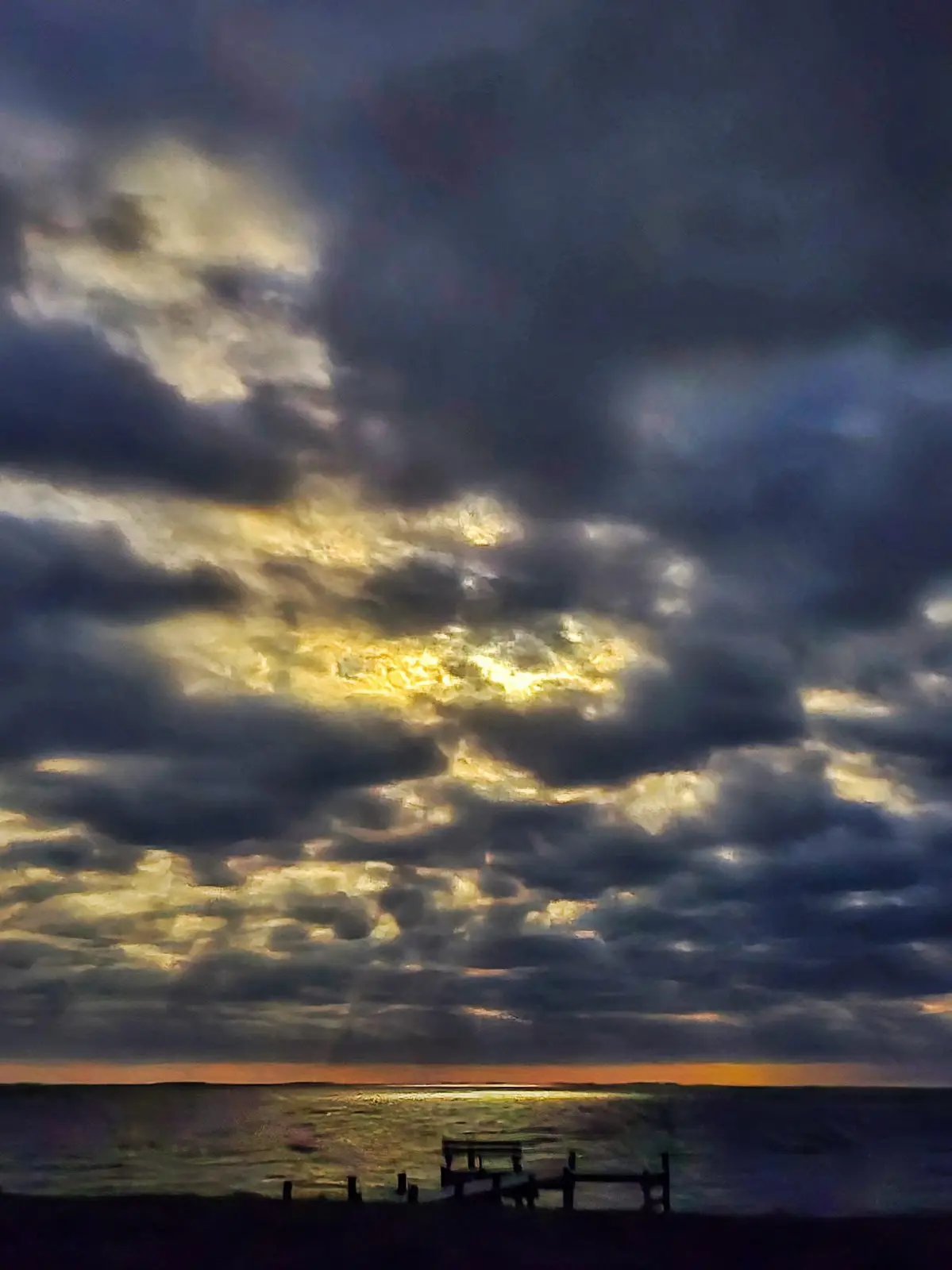 A dramatic, painterly sky with dark clouds illuminated by golden sunlight over a calm ocean, featuring a small wooden pier…