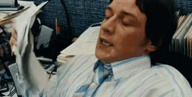 A man in a striped shirt and tie sits at a cluttered desk, visibly emotional as he holds up a white plastic bag. In the…