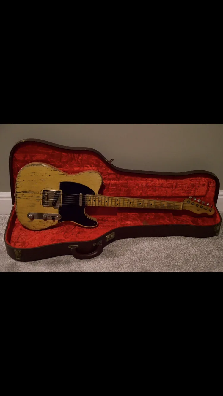 A heavily worn, light-wood-finish electric guitar with a black pickguard rests inside an open, red-lined hardshell case on a…