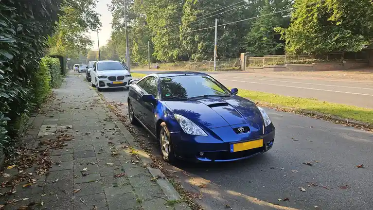 A blue Toyota sports car is parked on a street next to a sidewalk with fallen leaves, with a white BMW parked behind it. The…