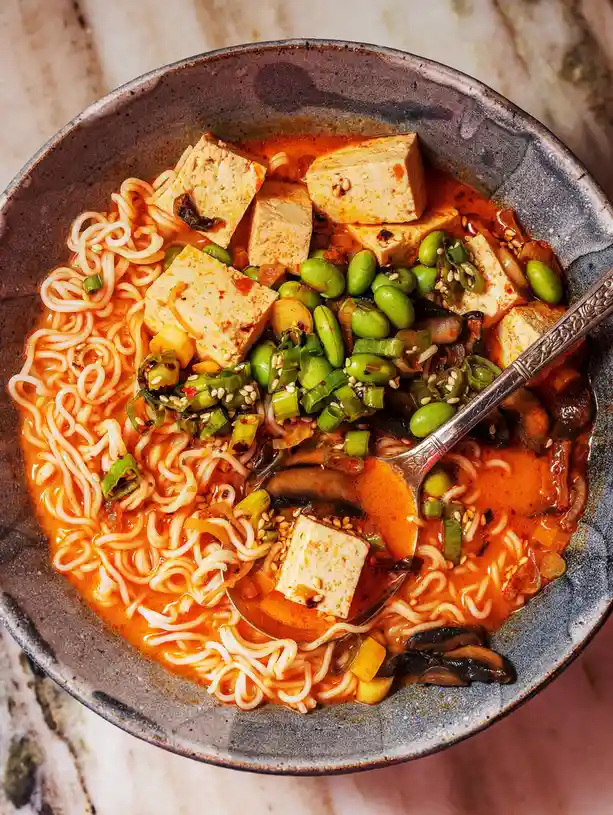 a top-down photo of a ceramic bowl filled with ramen noodles in a vibrant red broth, topped with cubed tofu, green edamame…
