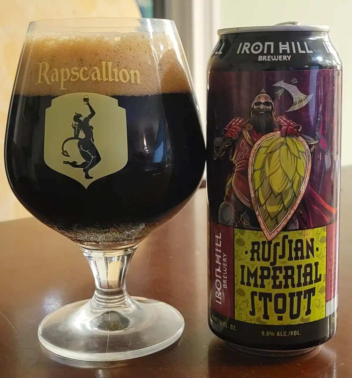 A dark beer in a stemmed glass labeled 'Rapscaillion' with a devilish logo sits next to a can of Iron Hill Brewery's…