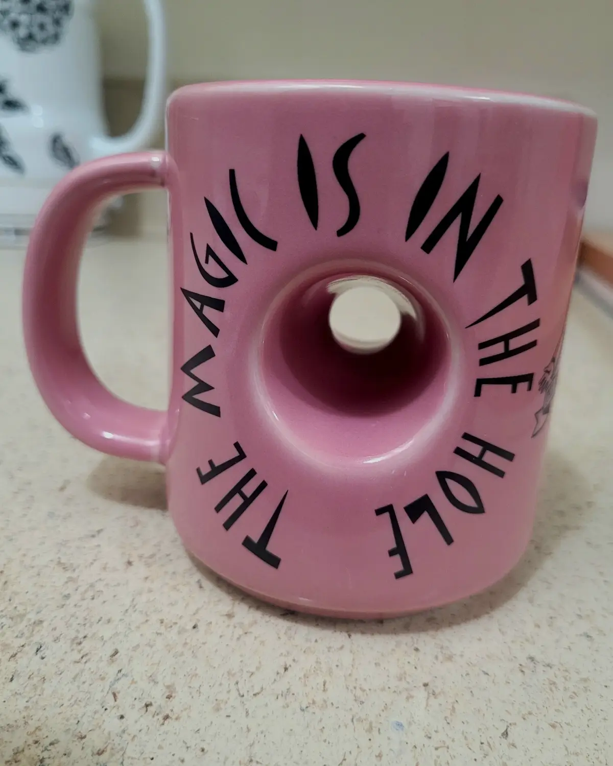 A pink ceramic mug shaped like a donut with a hole in the center, featuring the text 'THE MAGIC IS IN THE HOLE' printed in…