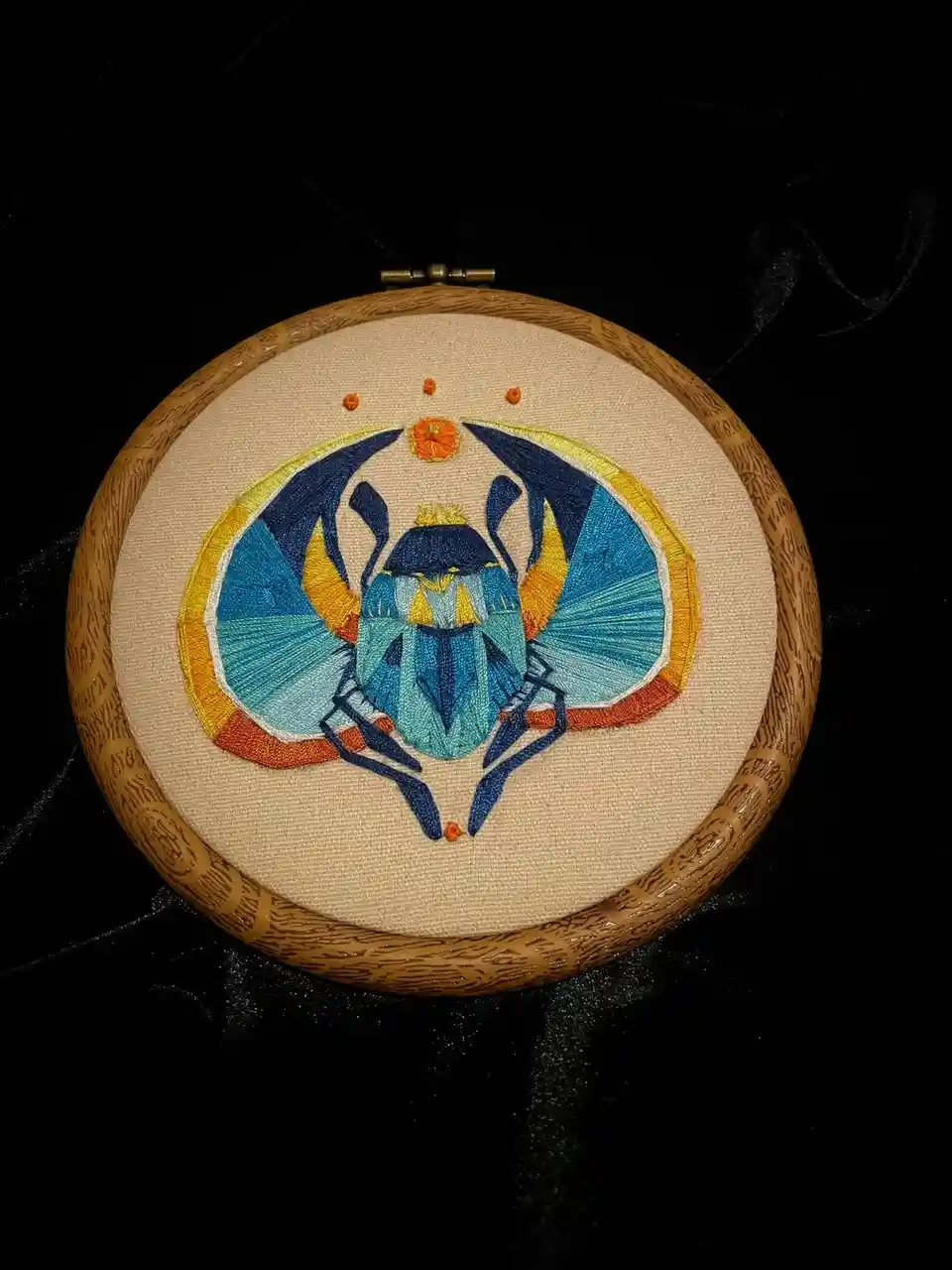 A circular embroidery hoop displays a colorful, stylized embroidered scarab beetle with blue, yellow, and orange threads…