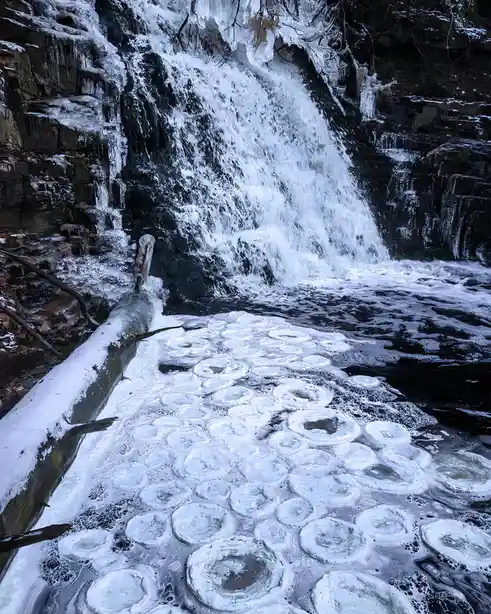 A winter scene shows a waterfall cascading over dark rocks, with a foreground of ice-covered water forming numerous…