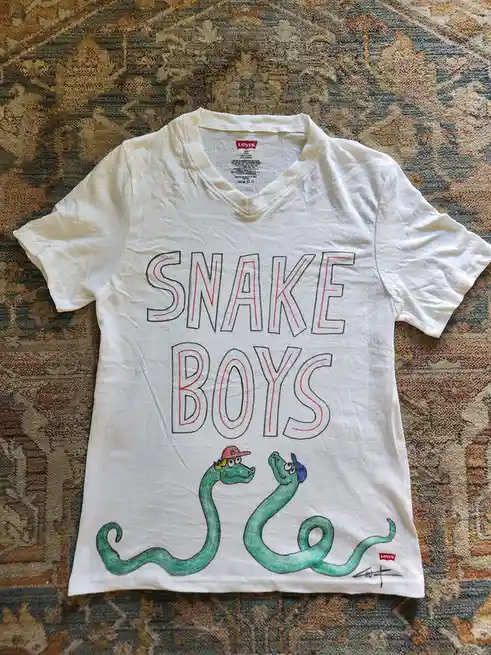 A white Levi's V-neck t-shirt laid flat on a patterned rug, featuring hand-drawn text 'SNAKE BOYS' in large outlined letters…